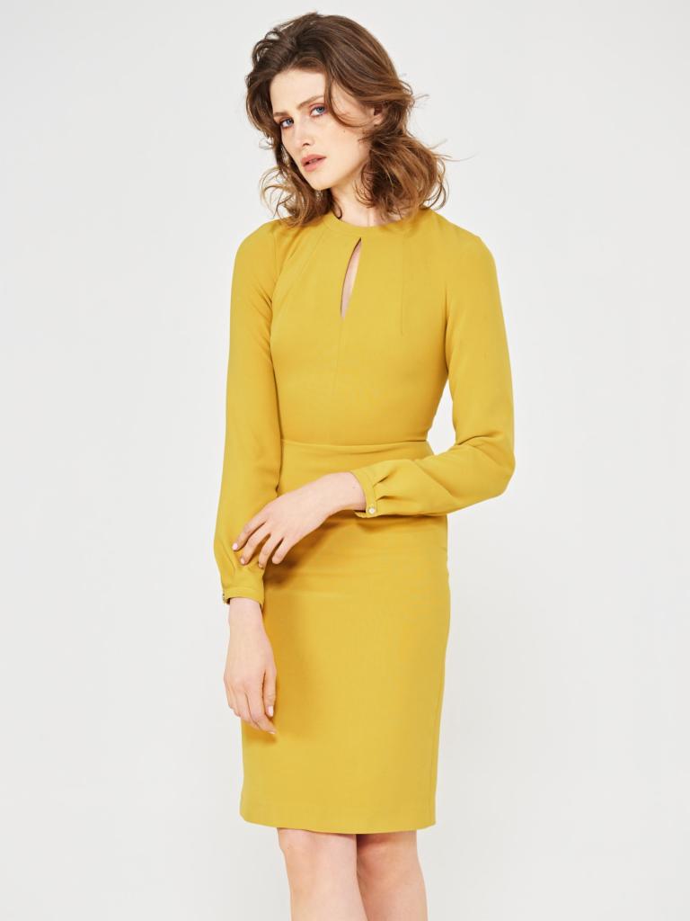 Kobieta prezentuje ołówkową sukienkę w odcieniu musztardowym z długimi rękawami i subtelnym wycięciem w dekolcie na białym tle.