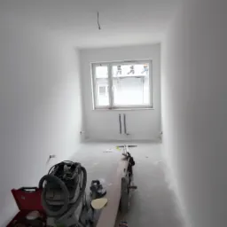 Wnętrze pokoju po nałożeniu gładzi, widoczne narzędzia: szlifierka do gładzi z odkurzaczem przemysłowym, szpachelki i pace, podłoga i ściany pokryte pyłem po szlifowaniu, okno w tle.