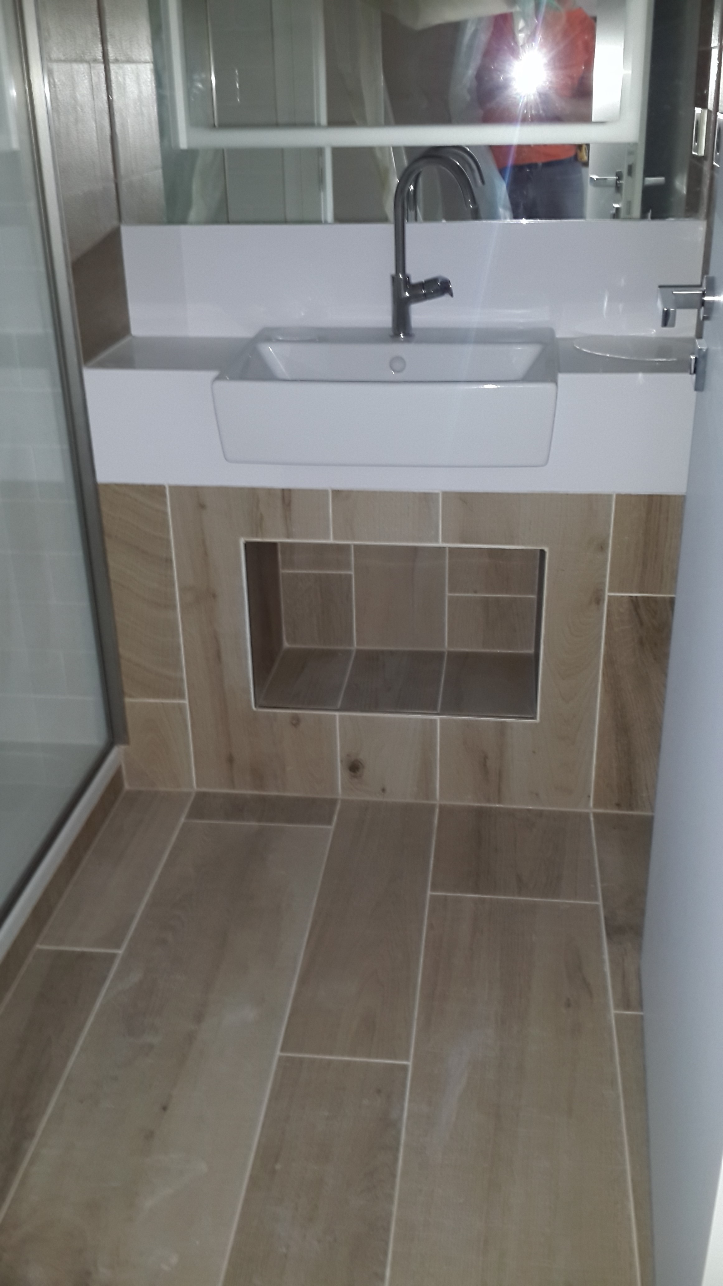 Nowoczesna łazienka po remoncie: biała umywalka nablatowa z chromowaną baterią, zabudowana płytkami imitującymi drewno, z wnęką pod umywalką. Widoczna kabina prysznicowa po lewej stronie i lustro...