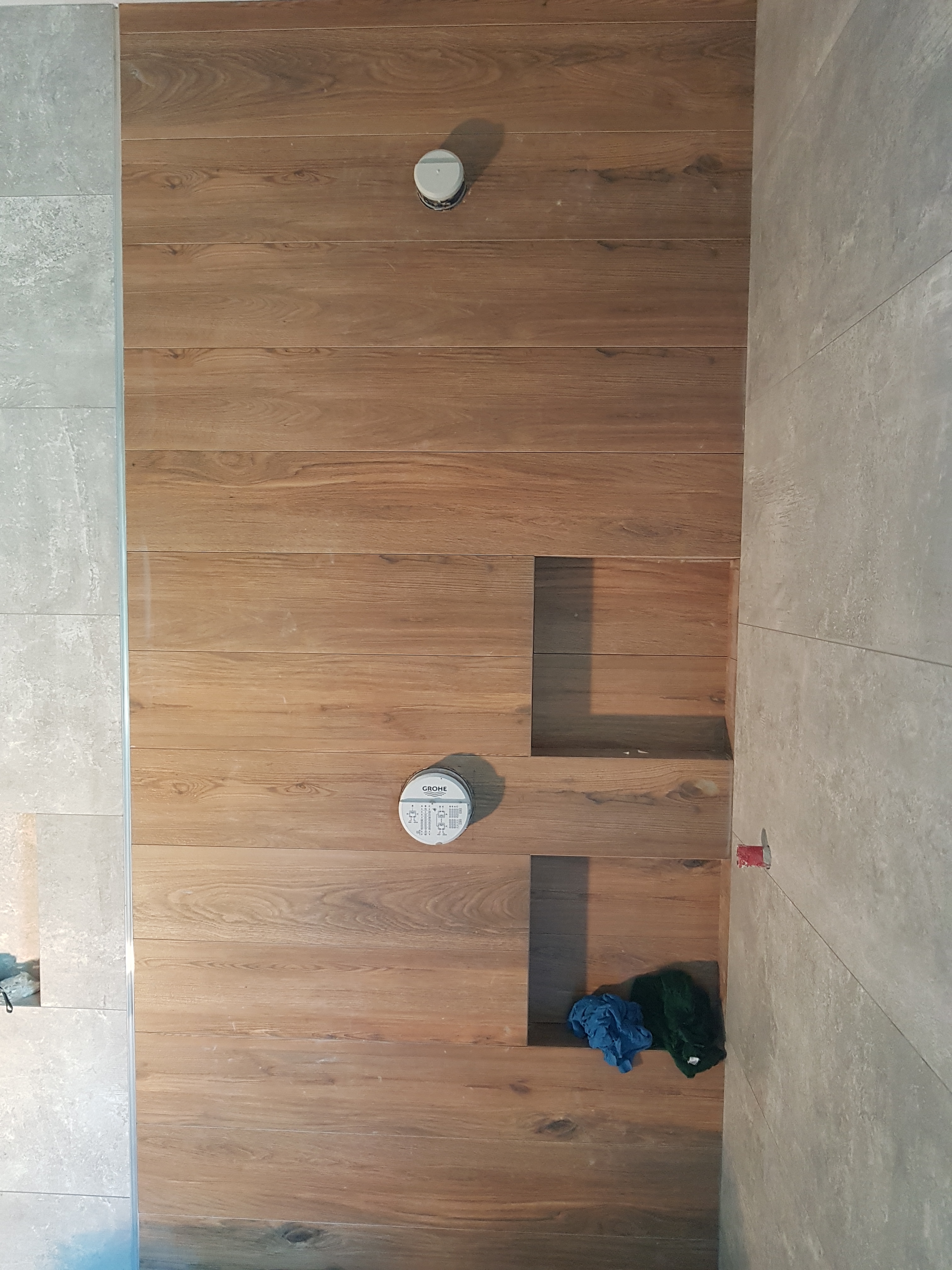 Ściana łazienki wyłożona płytkami imitującymi drewno, z widocznymi otworami na armaturę, obok ściana z szarych płytek. Na dolnej krawędzi ściany z drewnopodobnych płytek leżą niebieska i zielona...