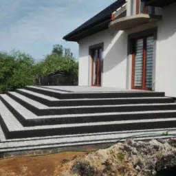 Schody wejściowe do domu wyłożone kostką granitową w kolorze szarym i czarnym, prowadzące na taras, z widocznym fragmentem elewacji budynku.