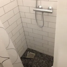 Narożnik kabiny prysznicowej wyłożony białymi, prostokątnymi płytkami i czarną mozaiką na podłodze, z widocznym odpływem i baterią prysznicową, zasłonięty częściowo wzorzystą zasłoną prysznicową.
