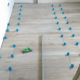 Proces układania jasnych płytek podłogowych imitujących drewno, z użyciem niebieskich klipsów poziomujących i szpachelki, widoczne narzędzia i materiały budowlane.