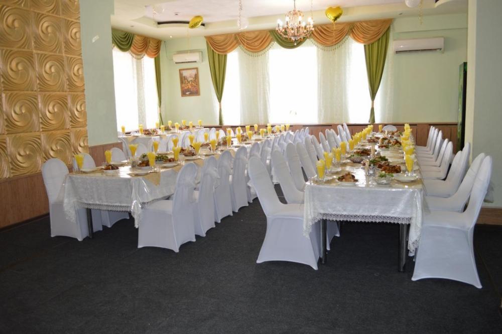 Uroczysta sala z długimi stołami nakrytymi białymi obrusami, krzesła w białych pokrowcach, na stołach dania i napoje w szklankach, złote balony w kształcie serc pod sufitem, dekoracyjne zasłony...