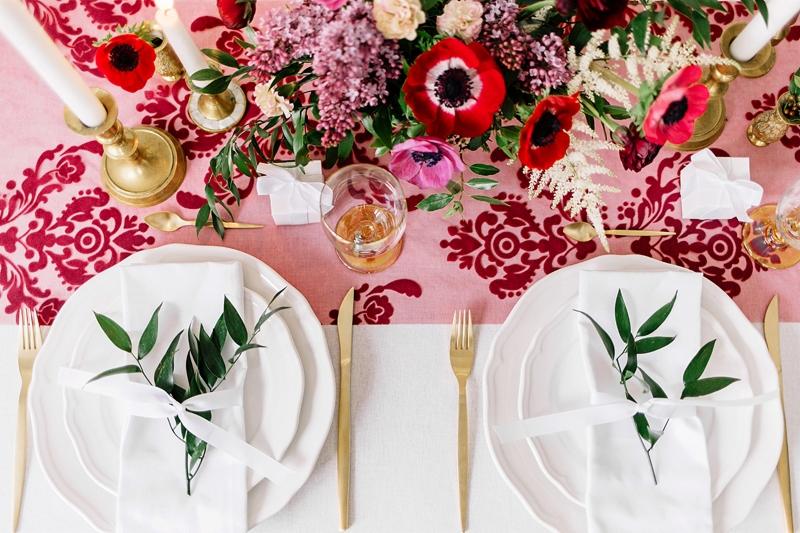 Elegancko nakryty stół z białymi talerzami, złotymi sztućcami, dekoracją z zielonych gałązek i białej wstążki na serwetkach, kwiatami w odcieniach różu i czerwieni oraz świecami w złotych...