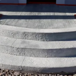 Betonowe schody wejściowe z efektem drewna na górnym podeście, zakończone kolumnami z pomarańczowymi podkładkami ochronnymi, otoczone drobnymi kamykami.