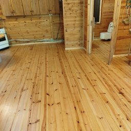 STYL-PARKIET DARIUSZ RODAK - Jasne, drewniane deski podłogowe w rustykalnym wnętrzu z boazerią. Widoczna kuchnia i łazienka w tle. Podłoga po renowacji, z widocznymi słojami drewna.