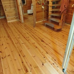 STYL-PARKIET DARIUSZ RODAK - Jasna, drewniana podłoga z widocznymi sękami w pokoju z drewnianymi ścianami i schodami. Widok na łazienkę w tle. Naturalne oświetlenie.