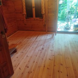 STYL-PARKIET DARIUSZ RODAK - Jasna, drewniana podłoga w domku z bali, widoczny fragment okna z zasłonami i drzwi. Na zewnątrz sprzęt do obróbki podłóg.