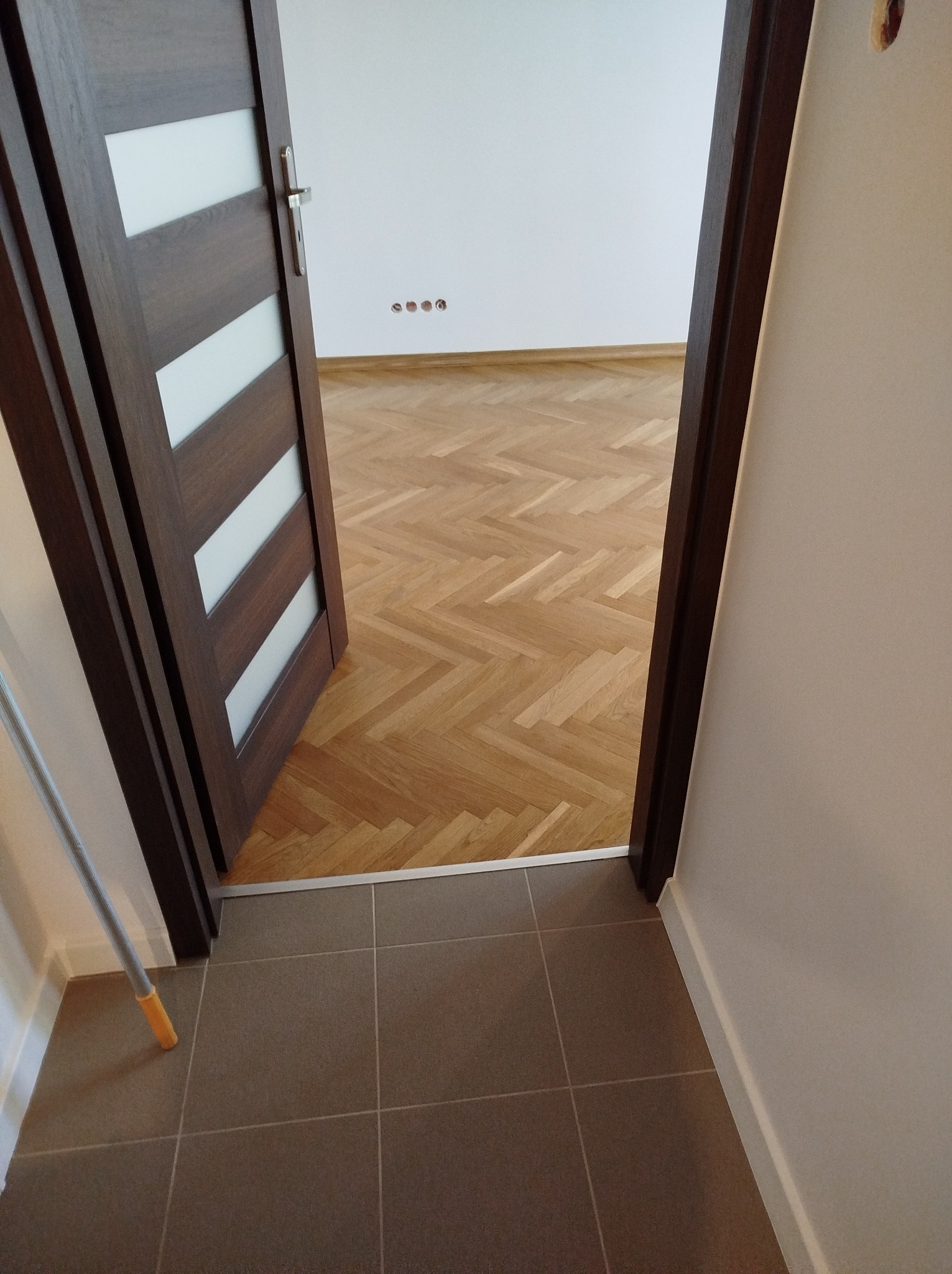 Widok na parkiet ułożony w jodełkę przez otwarte drzwi z brązowymi ramami i białymi wstawkami, widoczne gniazdka elektryczne w ścianie.