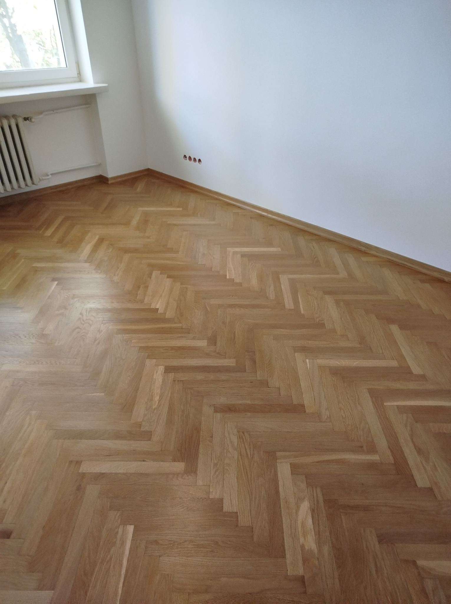 Ukończona podłoga z parkietu ułożonego w jodełkę klasyczną, widoczne listwy przypodłogowe i gniazdka elektryczne w ścianie.