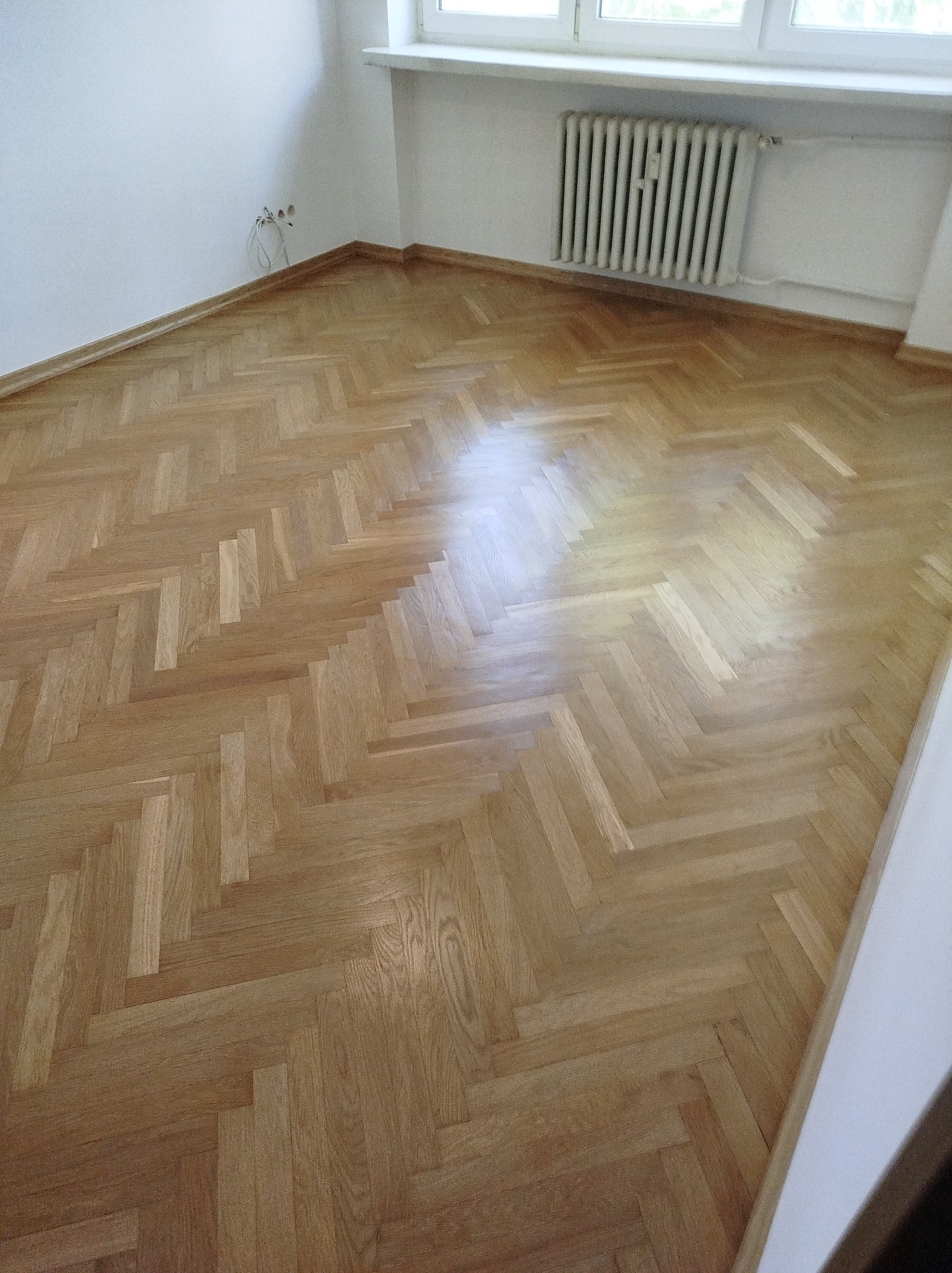 Jasny parkiet ułożony w jodełkę w pokoju z białymi ścianami, grzejnikiem i wystającymi kablami.
