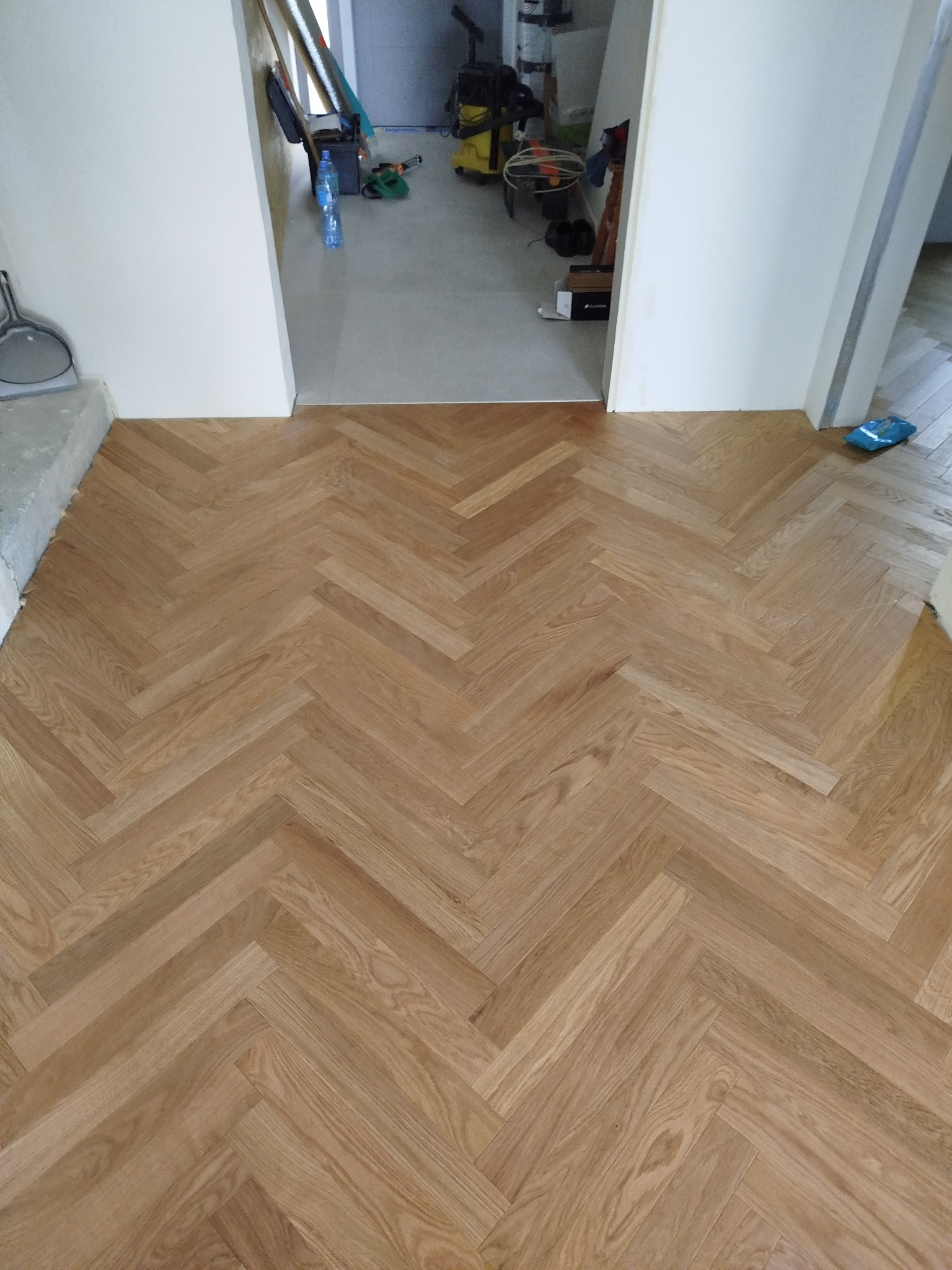 Jasny, dębowy parkiet ułożony we wzór jodełki francuskiej, widoczny z perspektywy wejścia do pomieszczenia. W tle widoczne narzędzia i materiały budowlane.