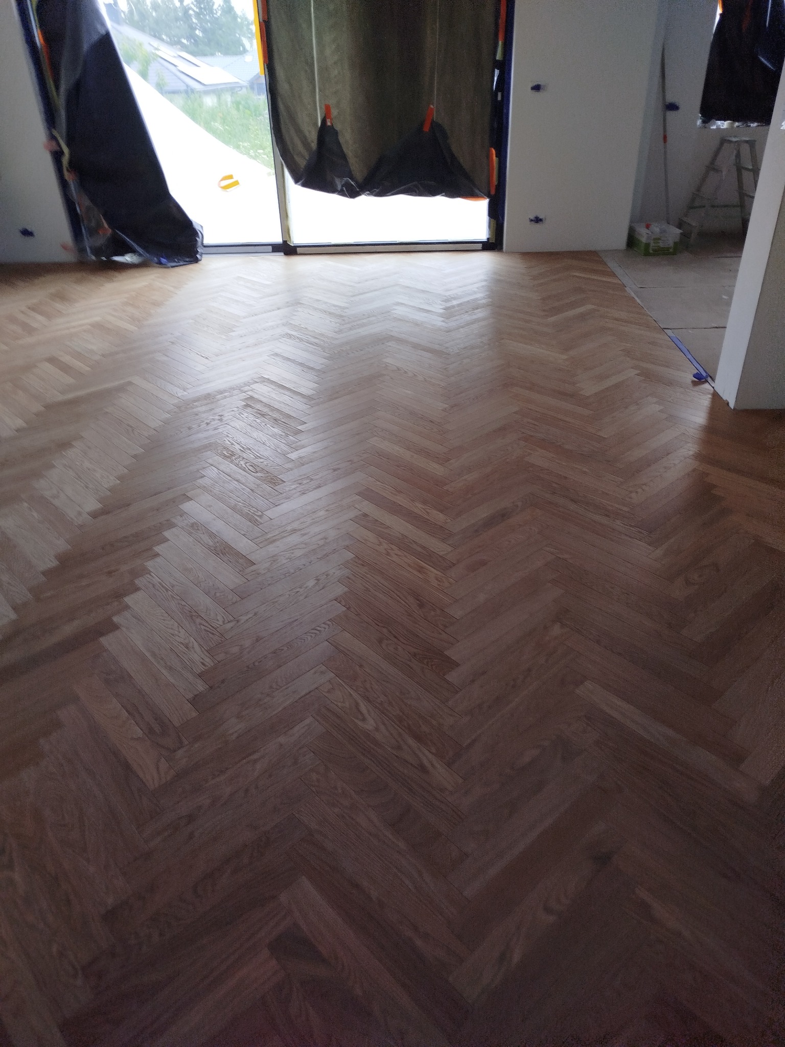 Parkiet ułożony w jodełkę na podłodze w jasnym, przestronnym pokoju. Okno zasłonięte folią ochronną. Widoczne resztki po remoncie.