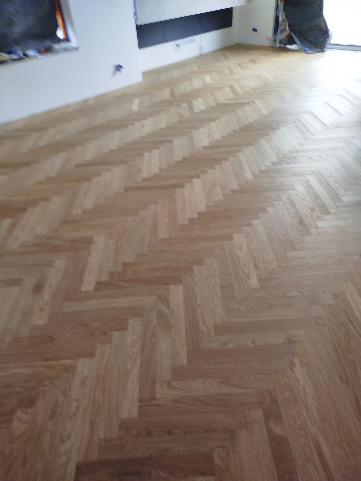 Ułożony parkiet w jodełkę francuską, jasne drewno, widok z góry, ściana z niebieskimi śladami po taśmie malarskiej w tle, naturalne światło.