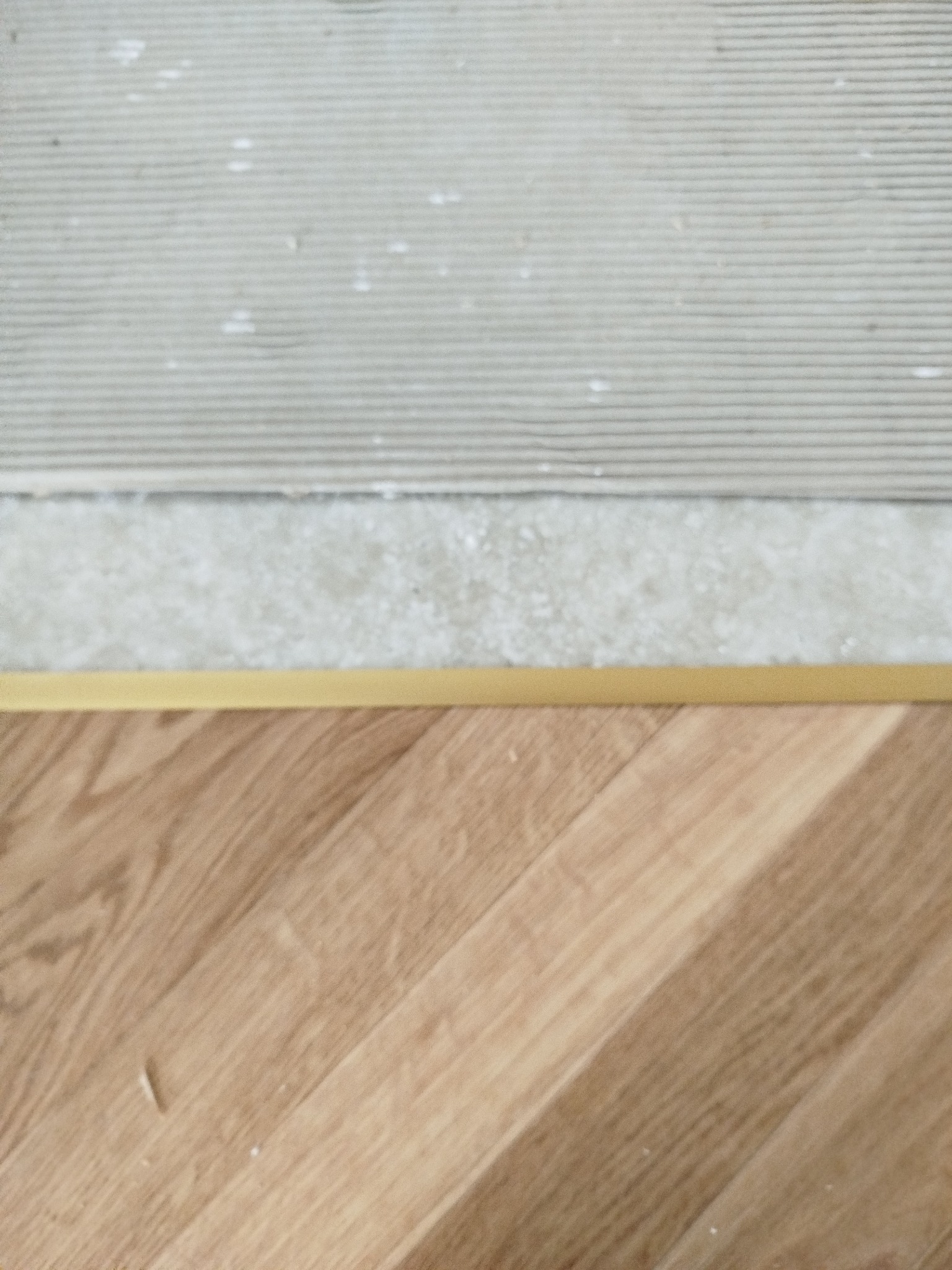 Fragment podłogi z parkietu ułożonego w jodełkę, oddzielonego złotą listwą od szarej, przygotowanej powierzchni pod dalsze prace wykończeniowe.