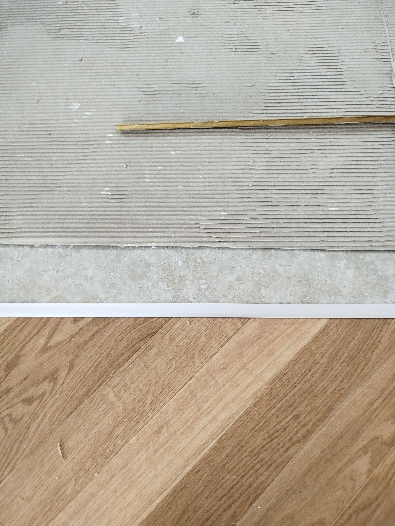Fragment podłogi: parkiet dębowy, listwa przypodłogowa i przygotowane podłoże z klejem. Widoczny detal łączenia różnych materiałów podłogowych.