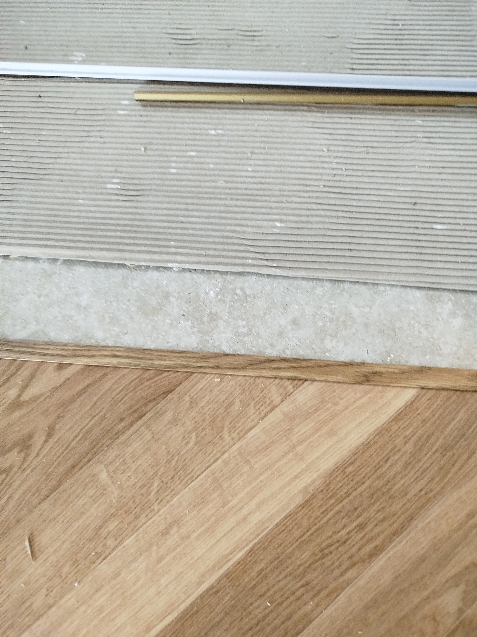 Fragment podłogi z ułożonym parkietem w jodełkę i listwą przypodłogową. Widoczne warstwy podkładu pod parkiet. Jasne drewno, ukośny wzór.