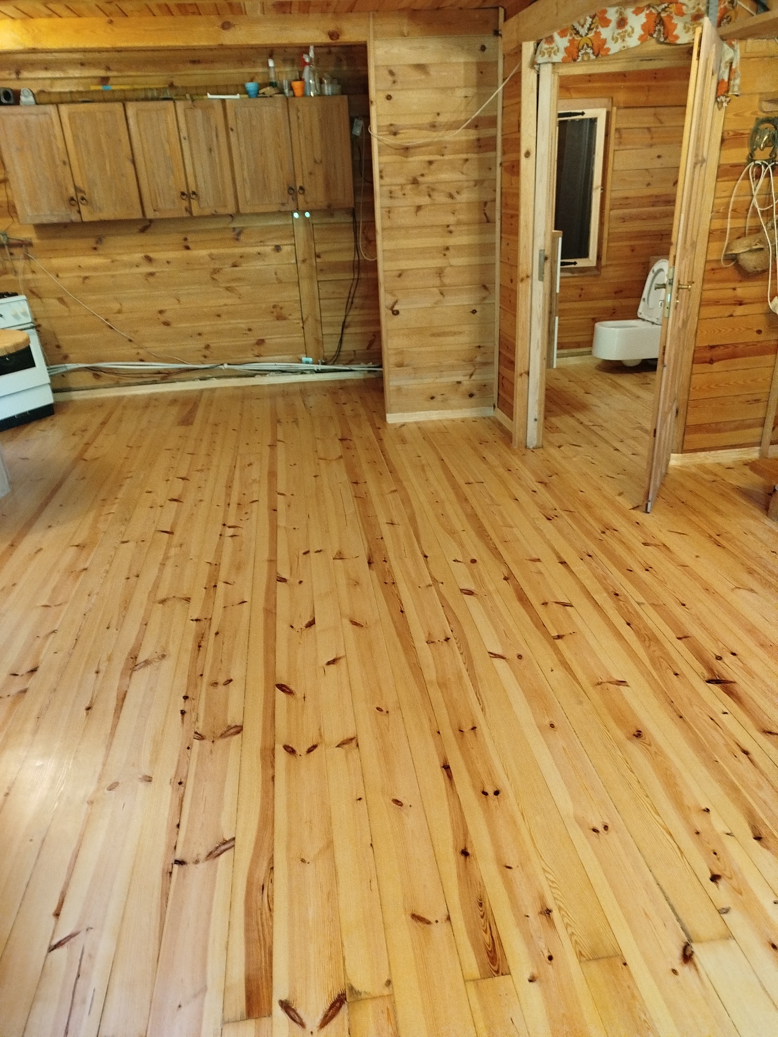 Jasne, drewniane deski podłogowe w rustykalnym wnętrzu z boazerią. Widoczna kuchnia i łazienka w tle. Podłoga po renowacji, z widocznymi słojami drewna.