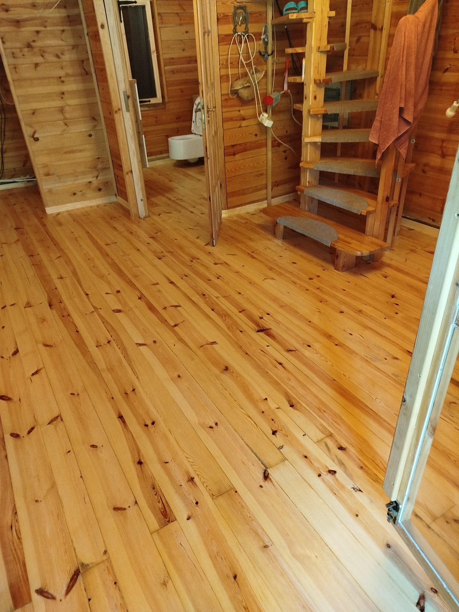 Jasna, drewniana podłoga z widocznymi sękami w pokoju z drewnianymi ścianami i schodami. Widok na łazienkę w tle. Naturalne oświetlenie.