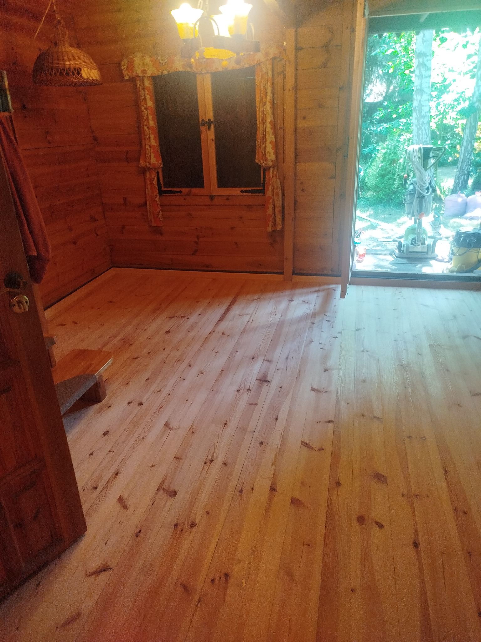 Jasna, drewniana podłoga w domku z bali, widoczny fragment okna z zasłonami i drzwi. Na zewnątrz sprzęt do obróbki podłóg.