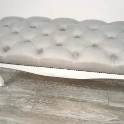 Szara tapicerowana pufa z pikowaniem typu chesterfield i białymi, giętymi nóżkami, wykończona ozdobnymi ćwiekami, na podłodze imitującej drewno.