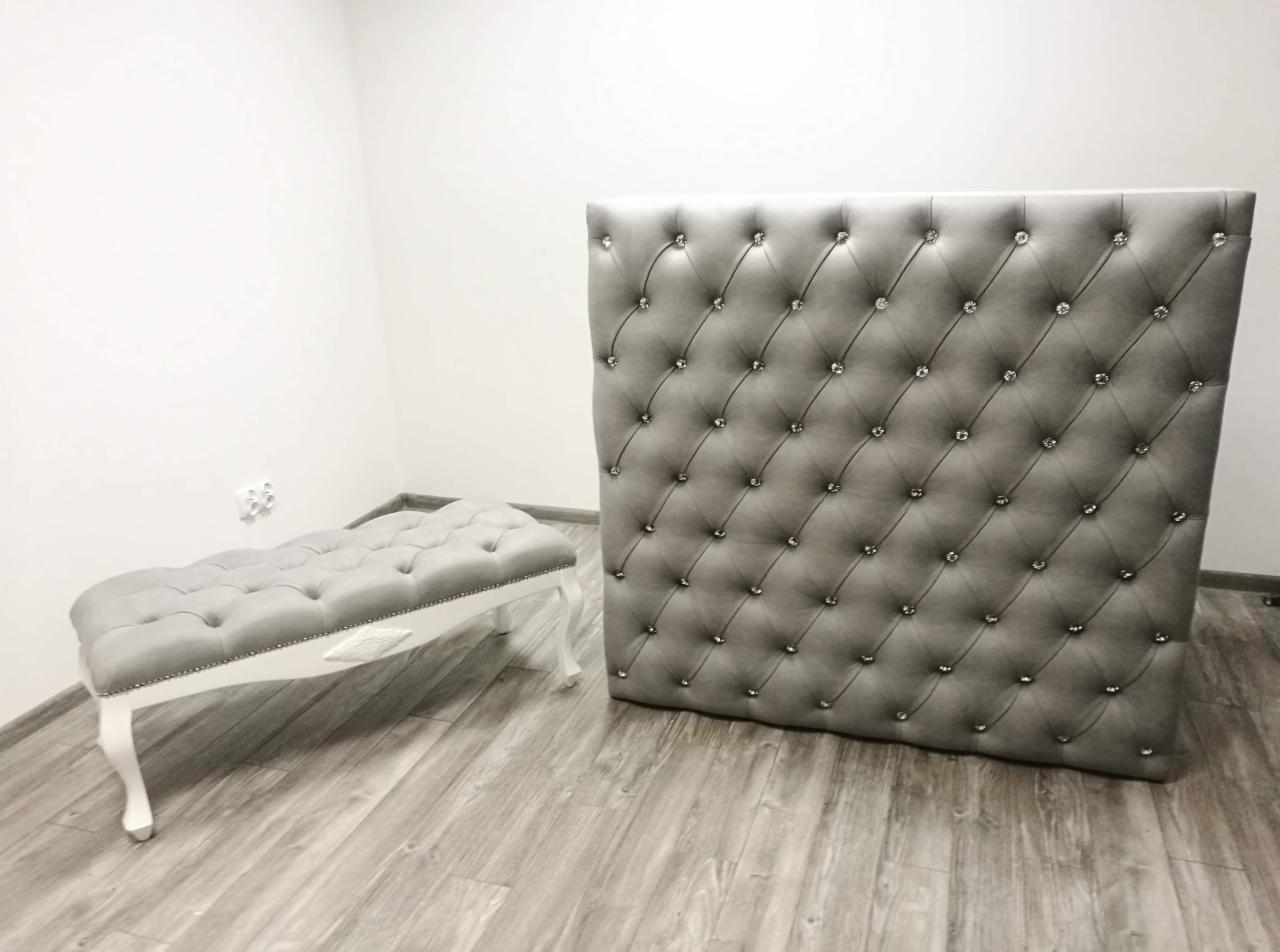 Szary, pikowany panel tapicerowany z kryształkami i dopasowana pufa na białych nóżkach w minimalistycznym wnętrzu.