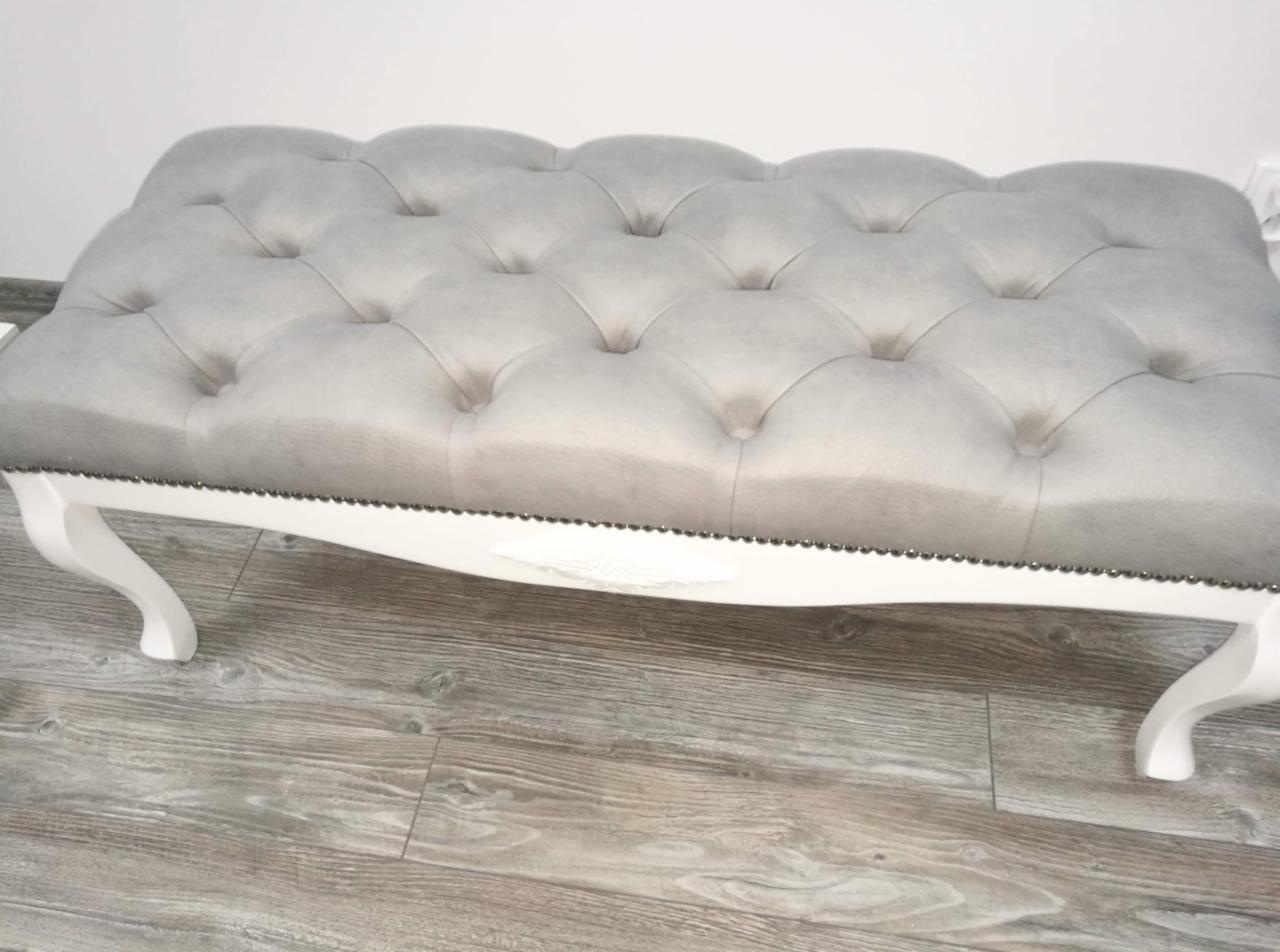 Szara tapicerowana pufa z pikowaniem typu chesterfield i białymi, giętymi nóżkami, wykończona ozdobnymi ćwiekami, na podłodze imitującej drewno.