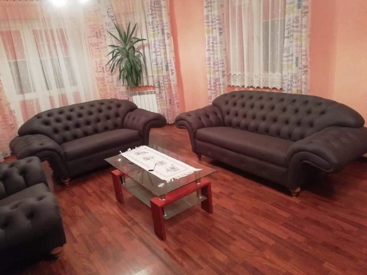 Dwie brązowe sofy Chesterfield z guzikami, fotel i stolik kawowy z szklanym blatem w salonie z parkietem i zasłonami w tle.