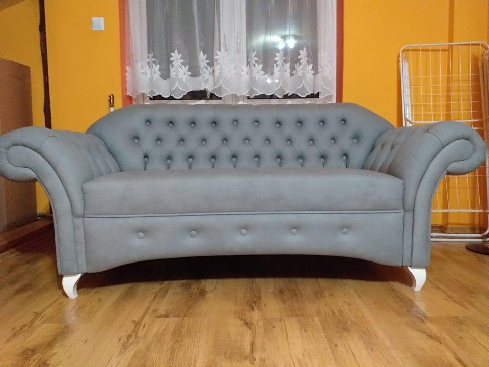 Szara sofa dwuosobowa w stylu Chesterfield z pikowanym oparciem i białymi nóżkami, ustawiona na drewnianej podłodze na tle żółtej ściany i okna z białą firanką.