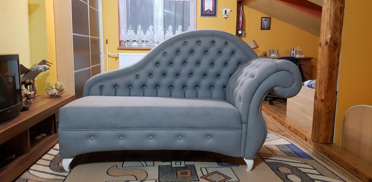 Szary, tapicerowany leżanka z pikowaniem w stylu Chesterfield, stojąca na dywanie w pokoju z żółtymi ścianami i drewnianymi elementami wykończenia.