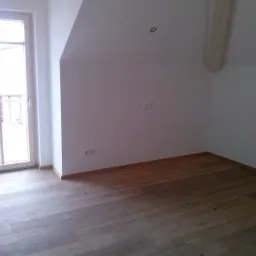 Puste, jasne poddasze z drewnianą podłogą, białymi ścianami i oknem balkonowym, widoczne gniazdka elektryczne.