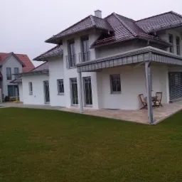 Nowoczesny, dwukondygnacyjny dom z białymi ścianami, ciemną dachówką i zadbanym trawnikiem, widziany z perspektywy ogrodu w pochmurny dzień.