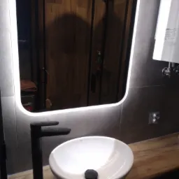 Nowoczesna łazienka z umywalką nablatową, czarną armaturą i podświetlanym lustrem LED, widoczna kabina prysznicowa z drewnopodobnymi płytkami w tle.