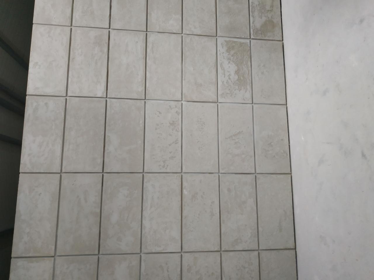 Ściana wyłożona szarymi, prostokątnymi płytkami z widocznymi smugami i niedoskonałościami, przylegająca do surowej, jasnoszarej ściany.