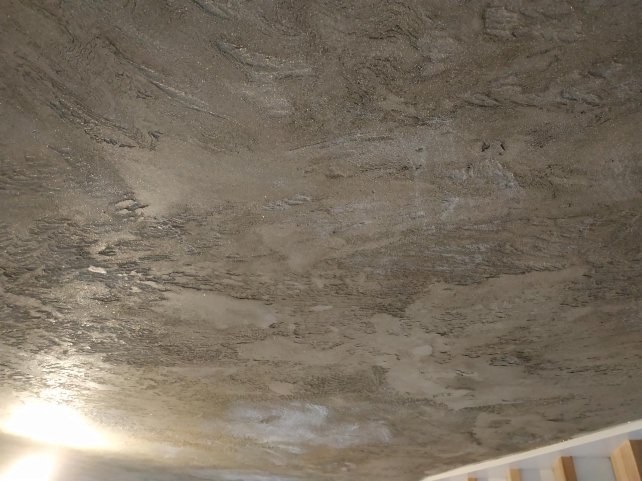 Teksturowany sufit z efektem betonu architektonicznego, widoczne nieregularne wzory i delikatne refleksy światła, wykończenie w odcieniach szarości i beżu.