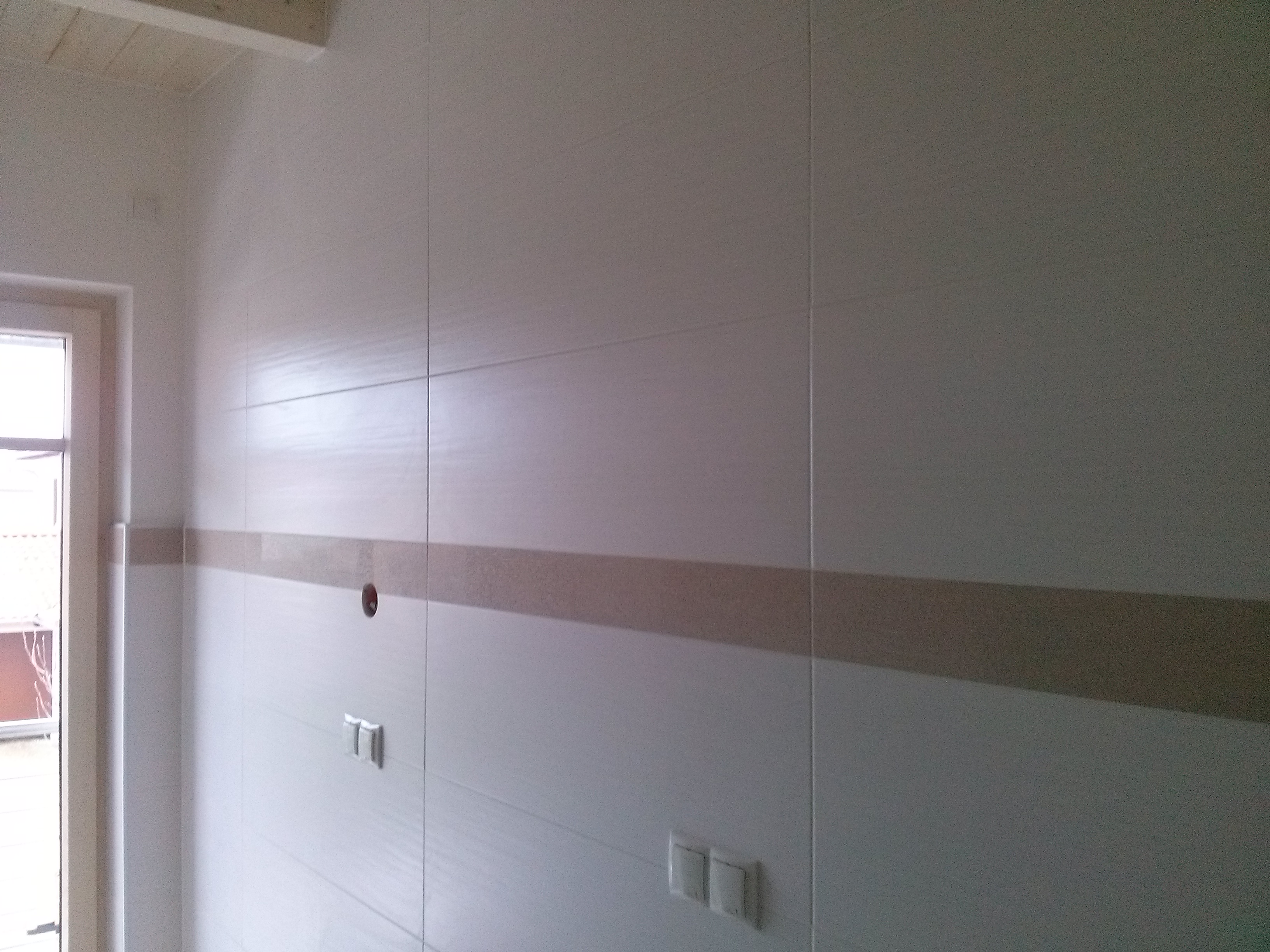 Ściana wyłożona jasnymi, prostokątnymi płytkami z poziomym pasem dekoracyjnym w odcieniach beżu, widoczne gniazdka elektryczne i otwór na rurę, fragment stolarki okiennej po lewej stronie.