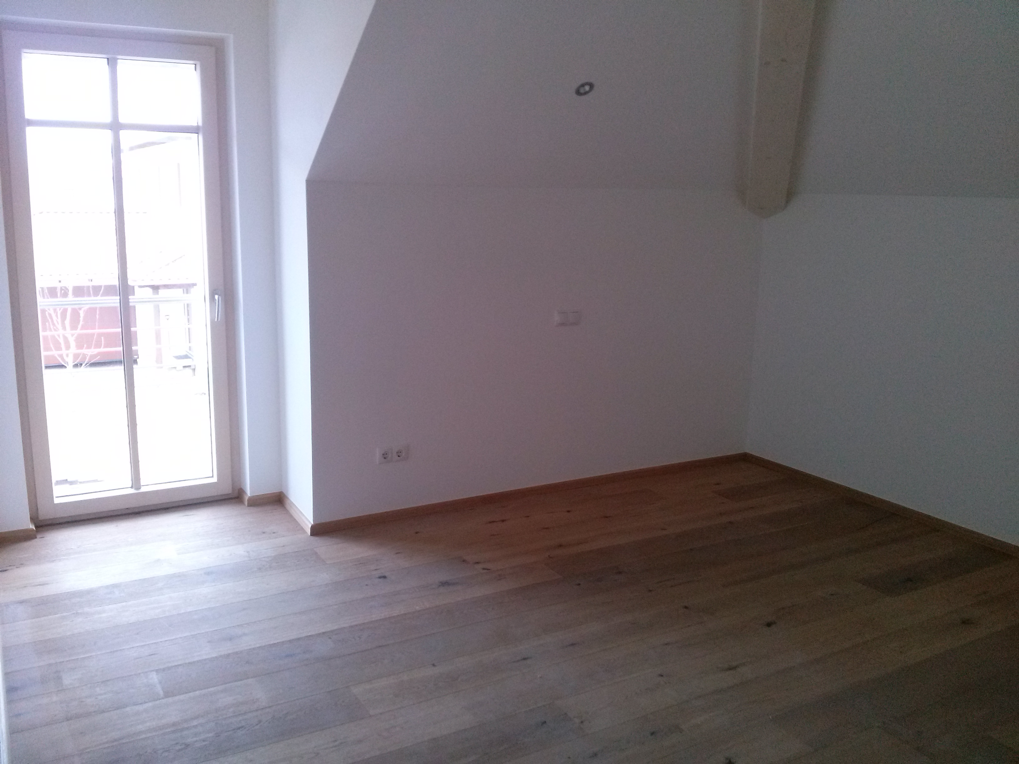 Puste, jasne poddasze z drewnianą podłogą, białymi ścianami i oknem balkonowym, widoczne gniazdka elektryczne.