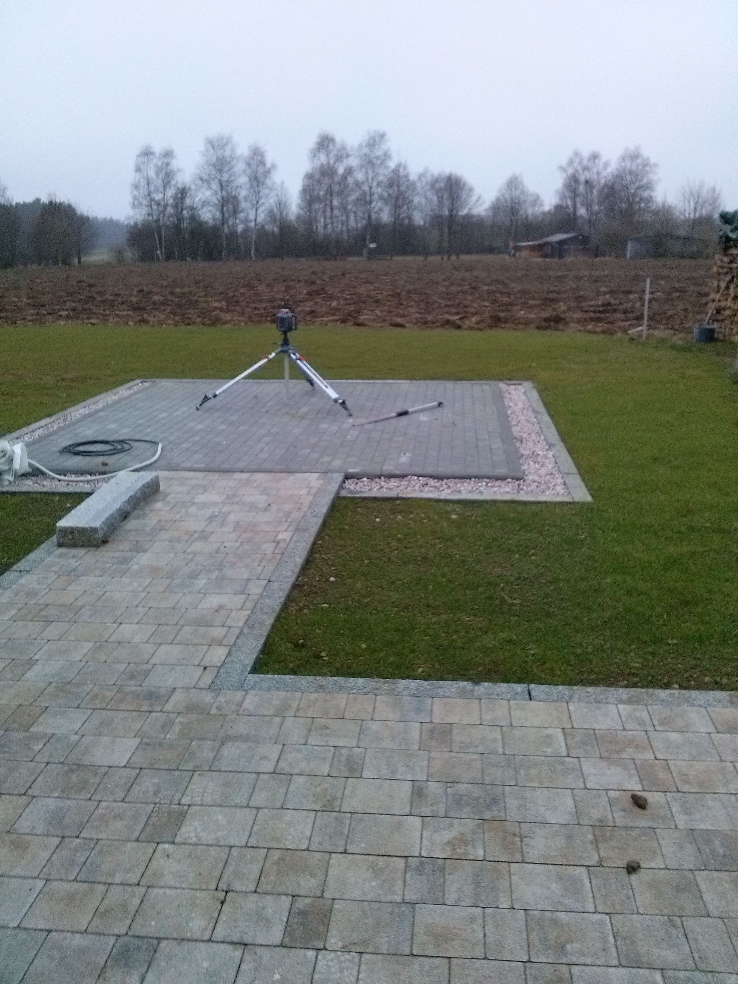 Ułożona kostka brukowa na posesji z widocznym niwelatorem laserowym na statywie, trawnikiem i polami w tle.