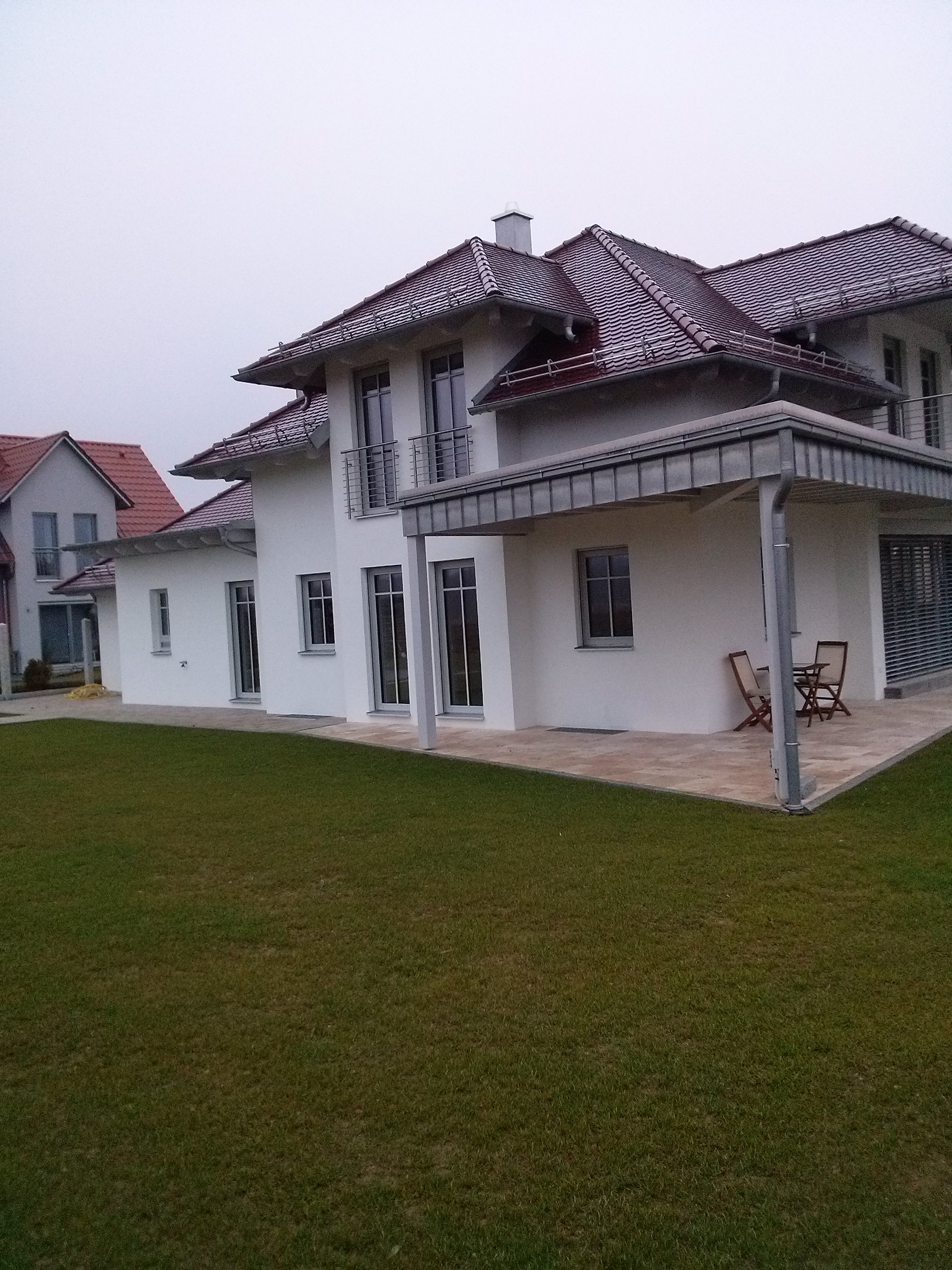 Nowoczesny, dwukondygnacyjny dom z białymi ścianami, ciemną dachówką i zadbanym trawnikiem, widziany z perspektywy ogrodu w pochmurny dzień.