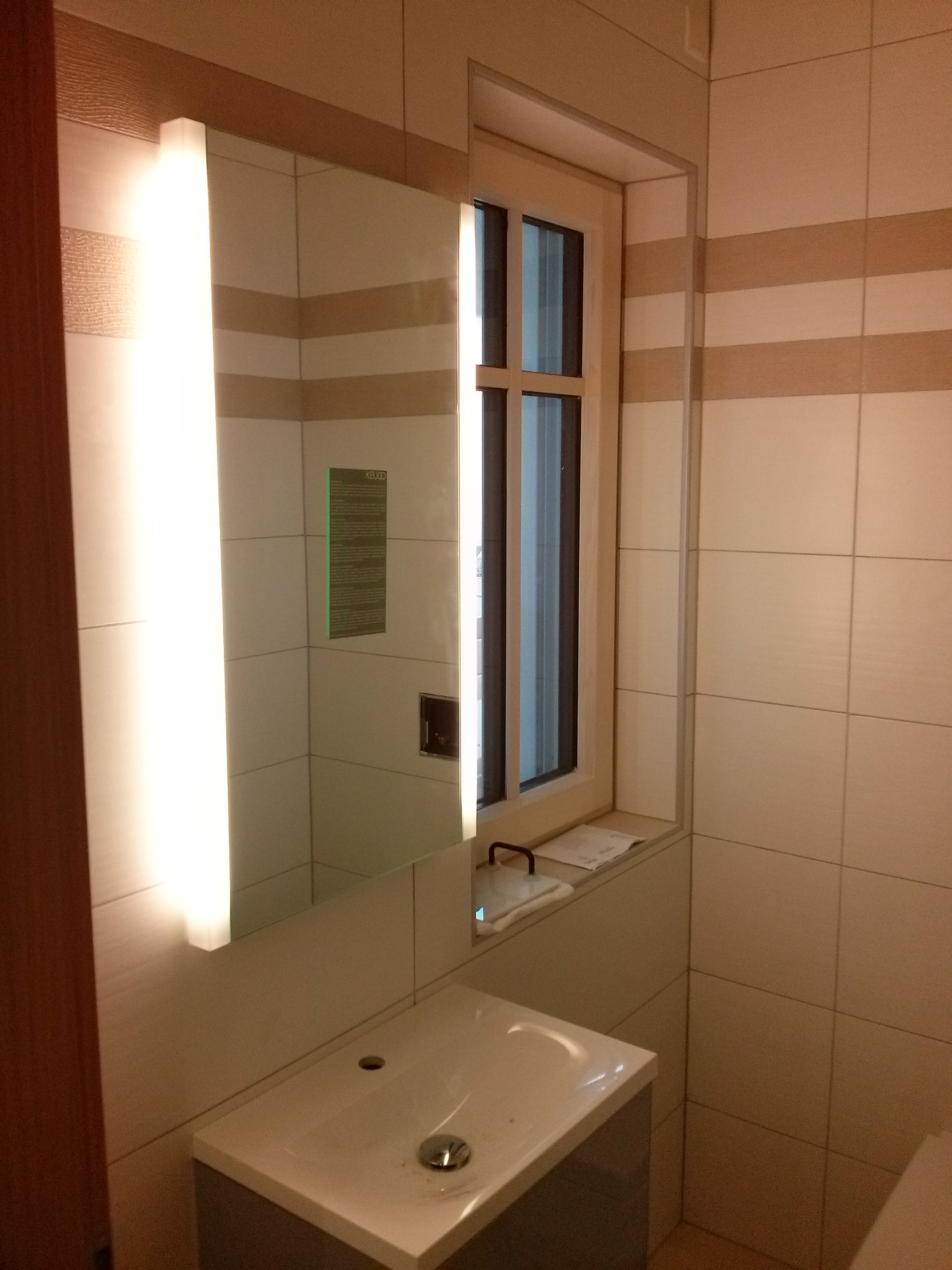 Nowoczesna łazienka z umywalką nablatową, podświetlanym lustrem z wbudowanym oświetleniem LED, oknem z drewnianą ramą i kwadratowymi płytkami w odcieniach beżu i bieli z poziomymi pasami.