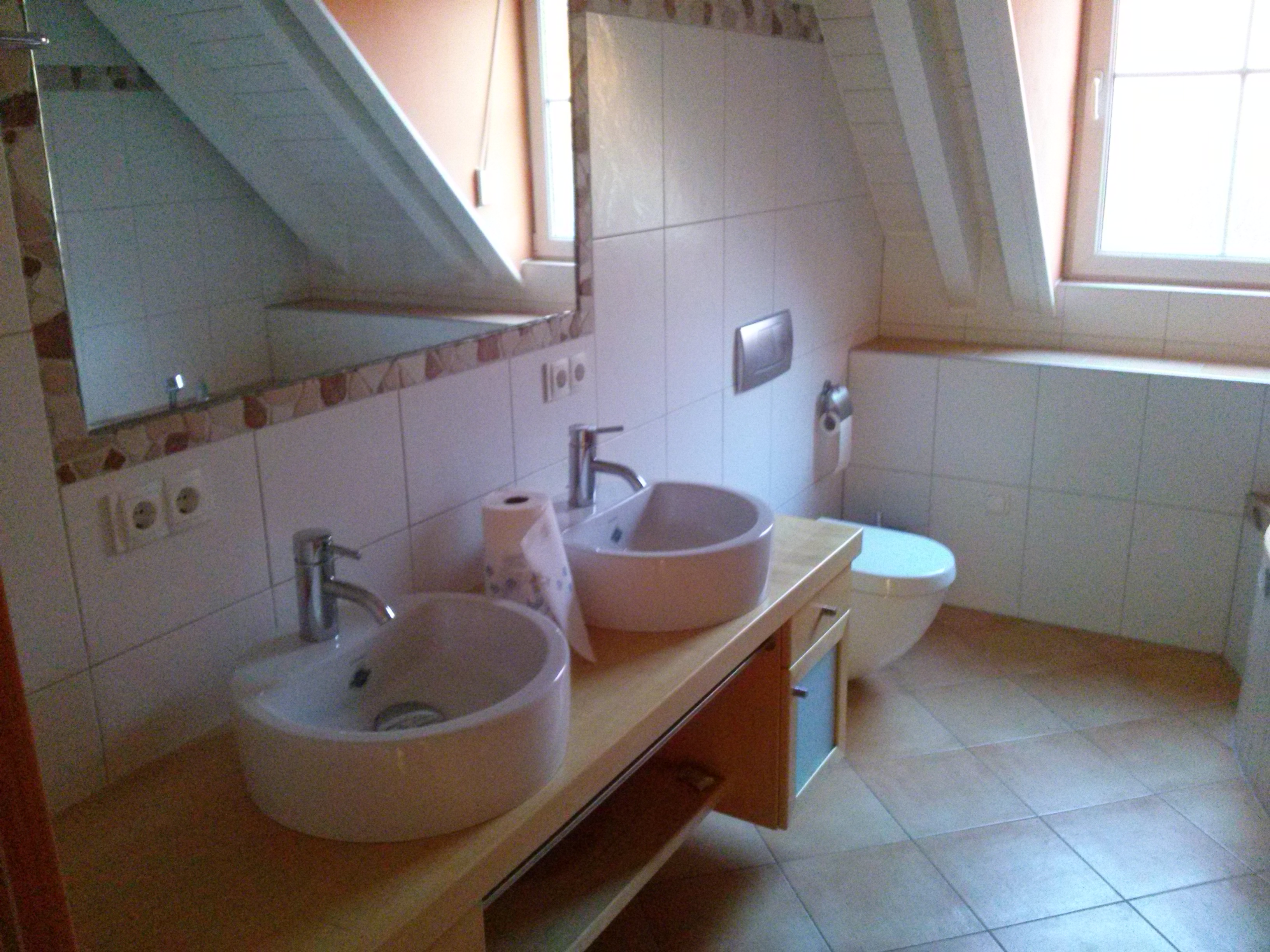 Jasna łazienka na poddaszu z dwoma okrągłymi umywalkami na drewnianym blacie, białe płytki na ścianach i podłodze, toaleta w tle.