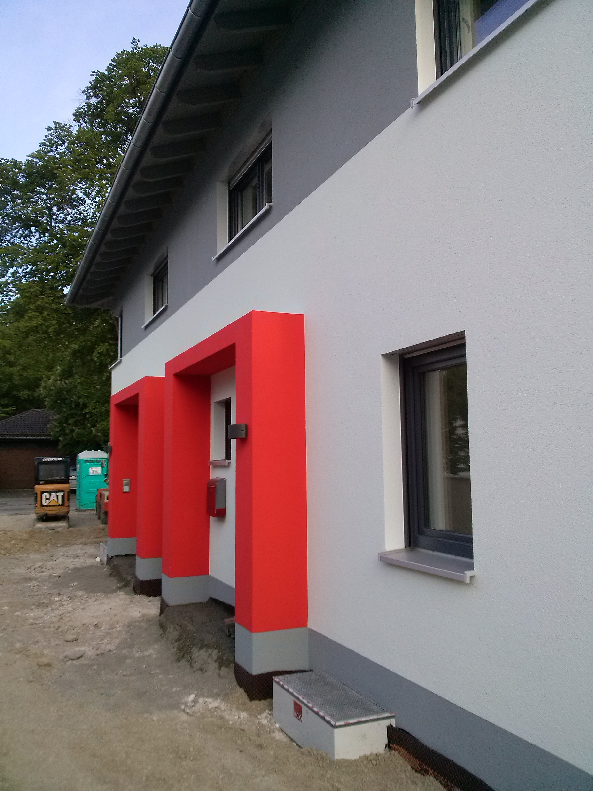 Dwukondygnacyjny budynek z elewacją w odcieniach szarości i bieli, z jaskrawoczerwonymi obramowaniami wejść, widoczny mały traktor i przenośna toaleta w tle.