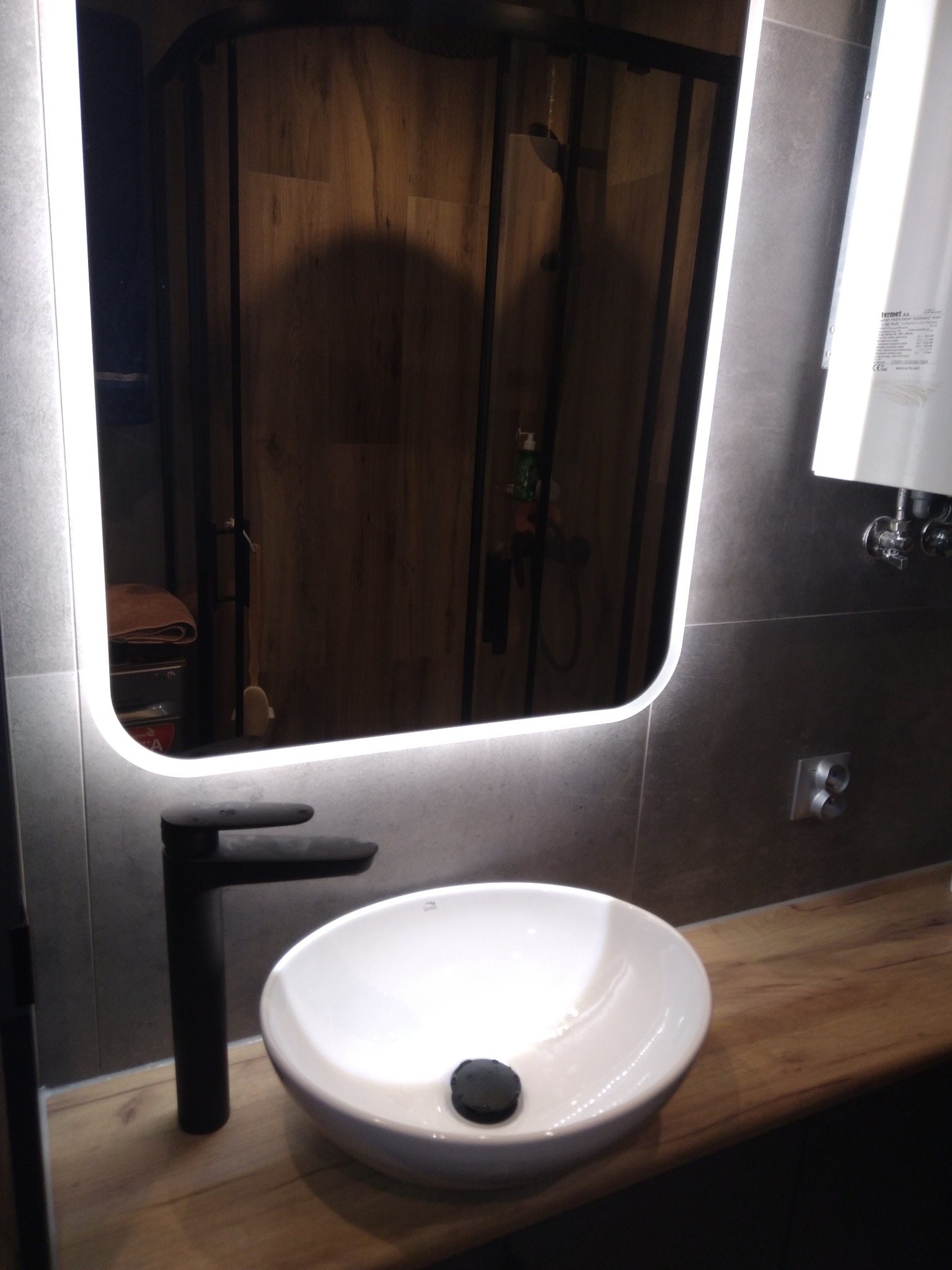 Nowoczesna łazienka z umywalką nablatową, czarną armaturą i podświetlanym lustrem LED, widoczna kabina prysznicowa z drewnopodobnymi płytkami w tle.