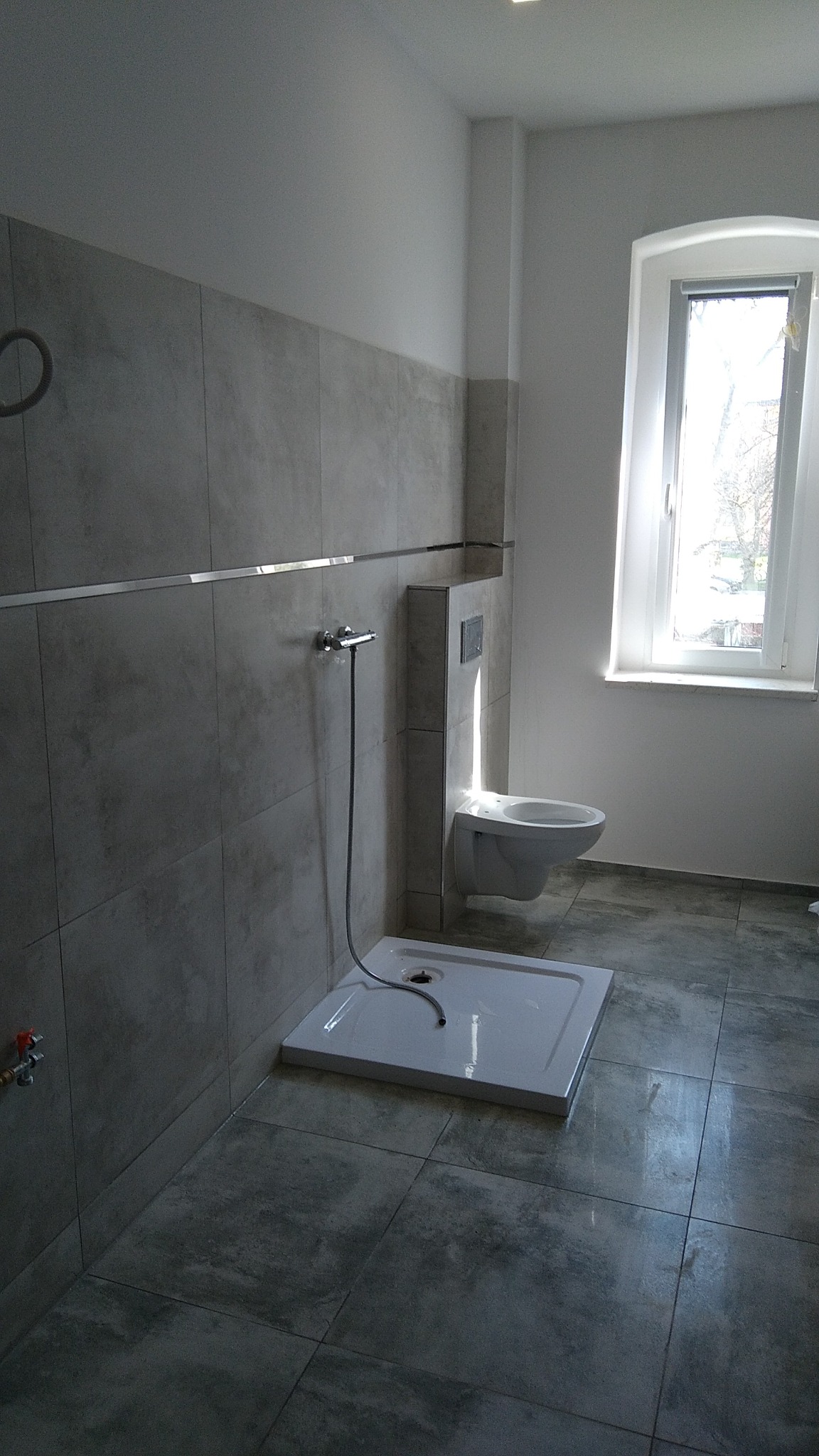 Nowa łazienka w odcieniach szarości z kabiną prysznicową bez brodzika, wiszącą toaletą i oknem z widokiem na drzewa.