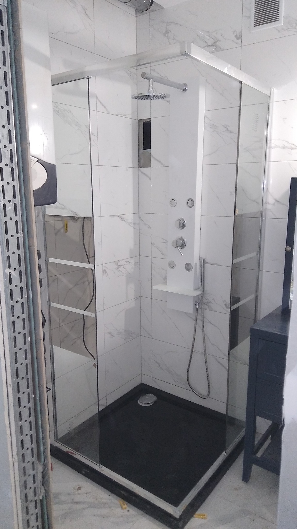 Kabina prysznicowa z hydromasażem i deszczownicą, wykończona białymi płytkami imitującymi marmur, czarny brodzik, częściowo widoczna szafka łazienkowa w kolorze granatowym.