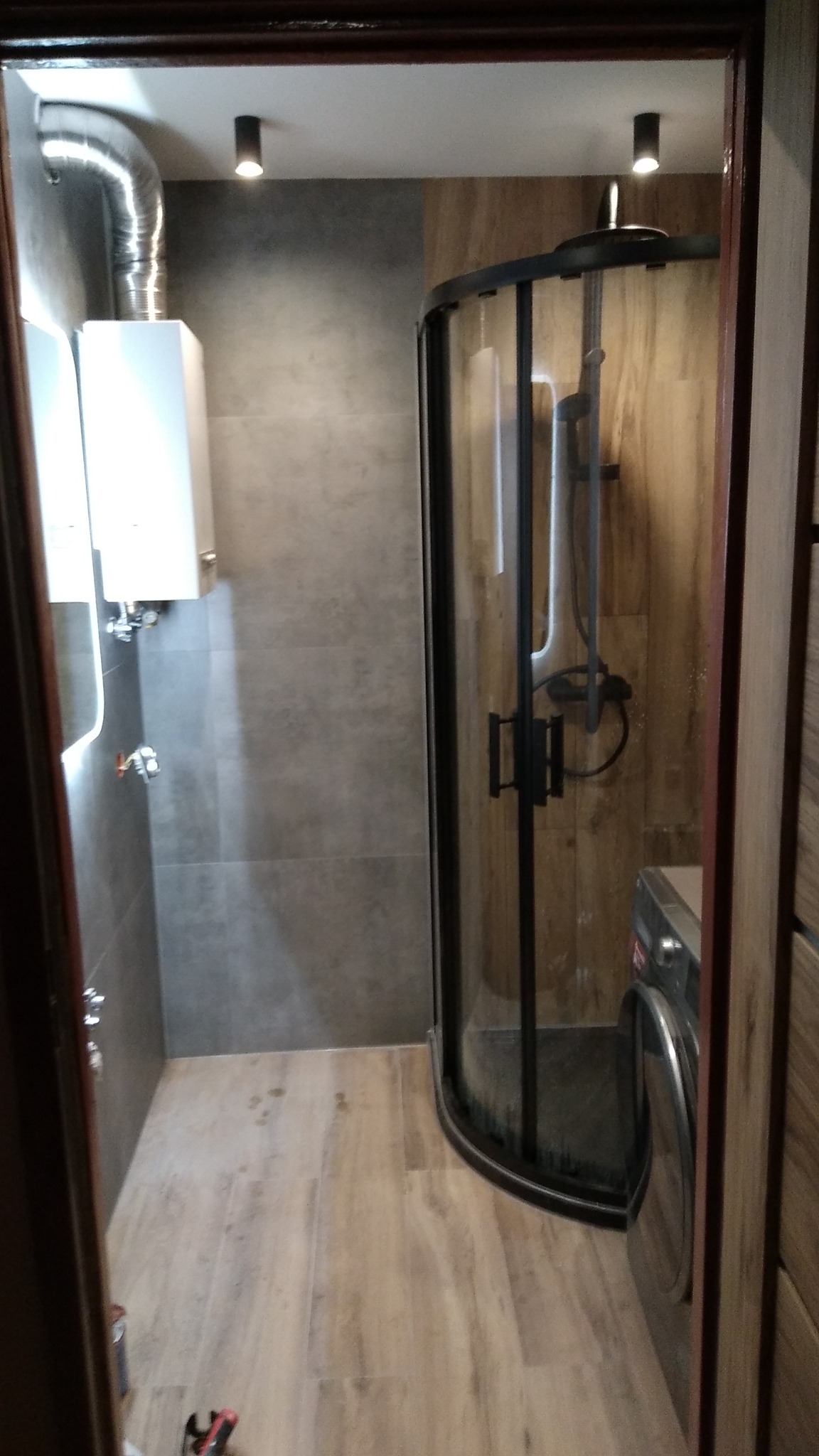 Nowoczesna łazienka z kabiną prysznicową z czarnymi profilami, płytki imitujące drewno na ścianie i podłodze, szara ściana z piecem gazowym i chromowaną rurą odprowadzającą spaliny.