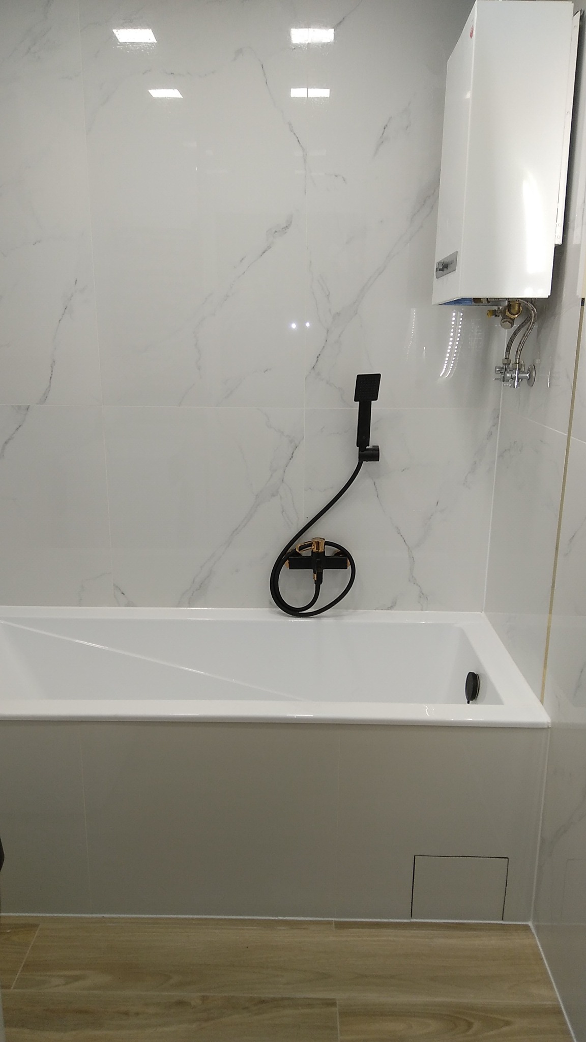 Biała wanna obudowana płytkami w łazience z marmurowymi kaflami, czarna bateria bidetowa i biały podgrzewacz wody wiszący na ścianie.