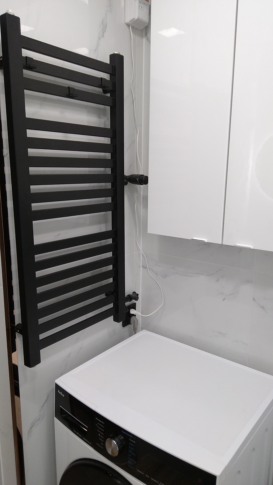 Czarny, drabinkowy grzejnik łazienkowy na tle marmurowej ściany, obok biała pralka Amica i wisząca szafka. Widoczne elementy instalacji hydraulicznej i elektrycznej.