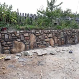 Wznoszenie kamiennego muru oporowego na nierównym terenie, widoczny poziomica i spawarka, w tle drewniane ogrodzenie i zieleń.